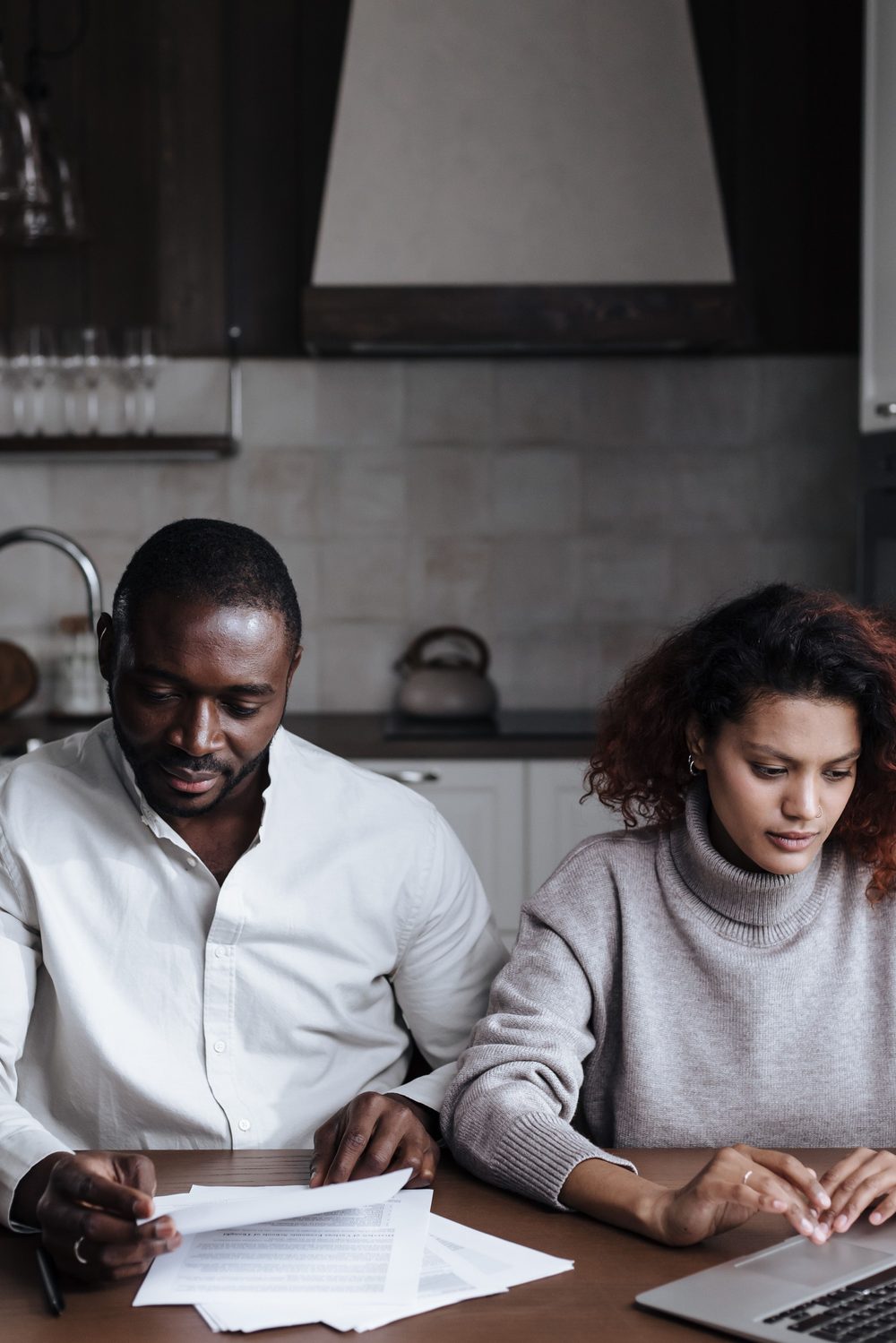 A couple sitting together at their kitchen table, reviewing paperwork with a laptop open between them.