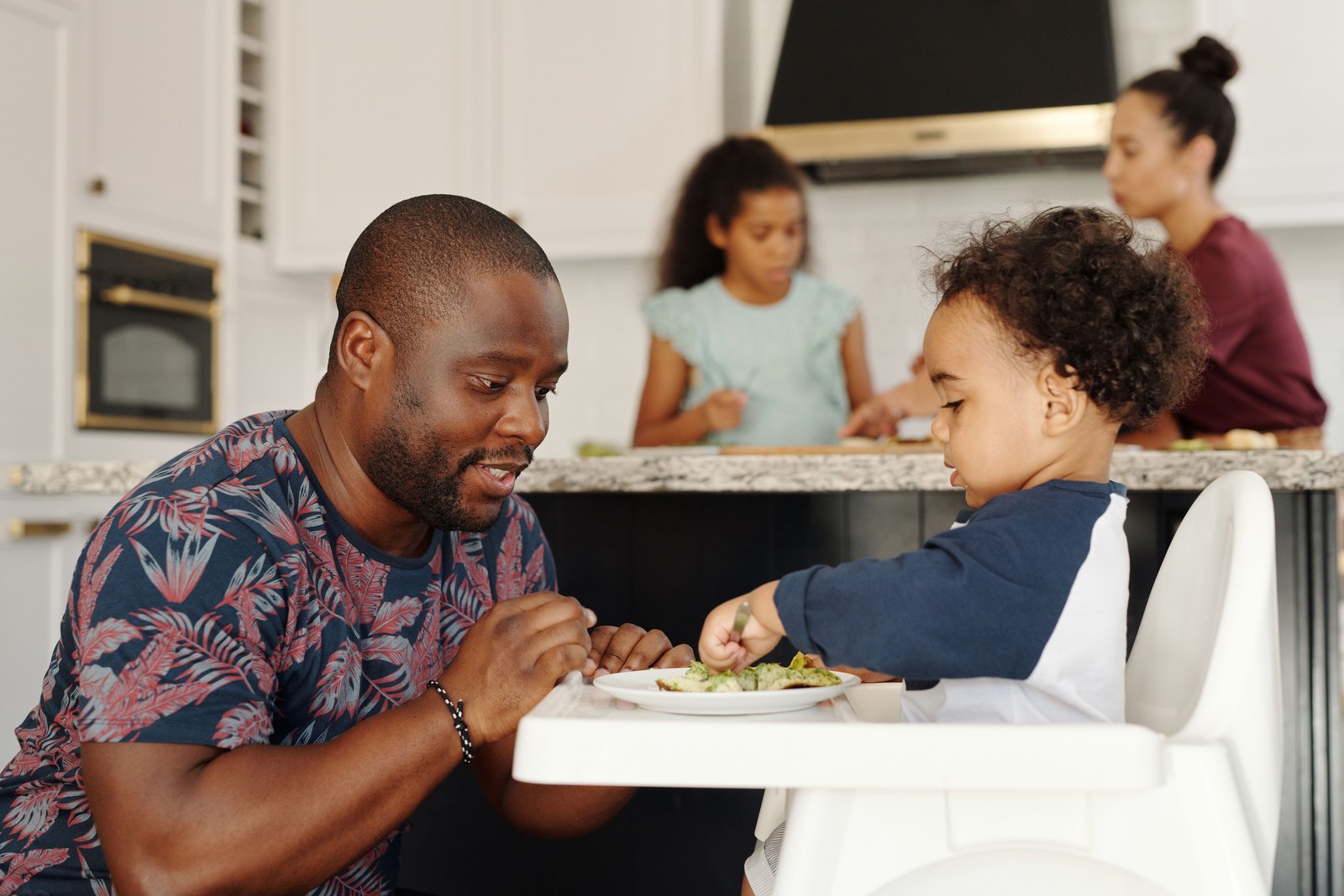A father sitting beside his toddler's high chair at breakfast while the rest of the family prepares food at the kitchen island behind them.